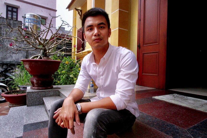 Hoang Vu Duy, with black hair and a lilac shirt, sits outside a yellow Vietnamese house with small garden.
