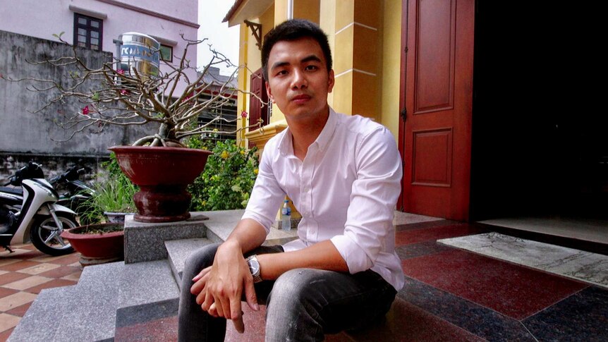 Hoang Vu Duy, with black hair and a lilac shirt, sits outside a yellow Vietnamese house with small garden.