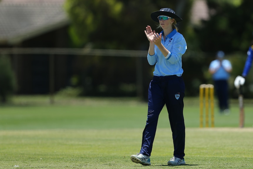 Woman standing on cricket pitch, clapping, wearing blue and navy uniform and wide brim hat