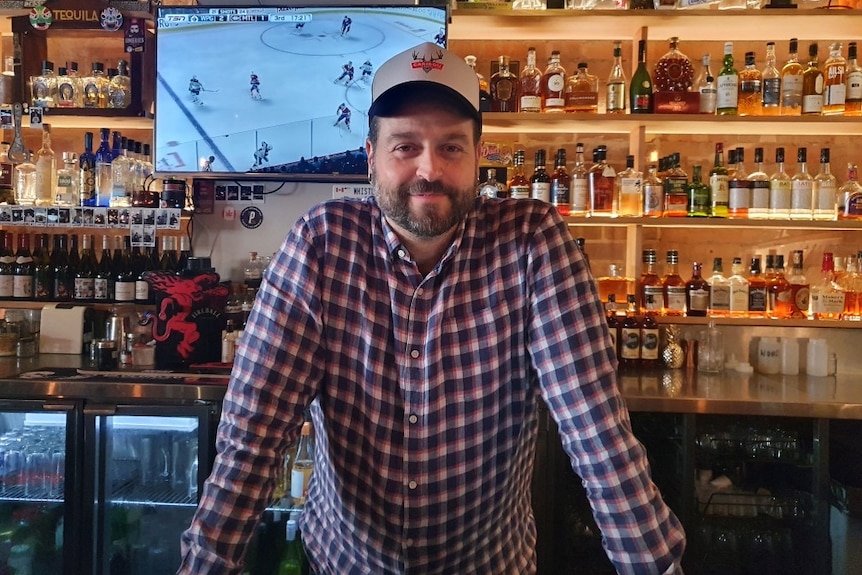 A man stands in front of a bar. Ice hockey is being played on a TV in the background.