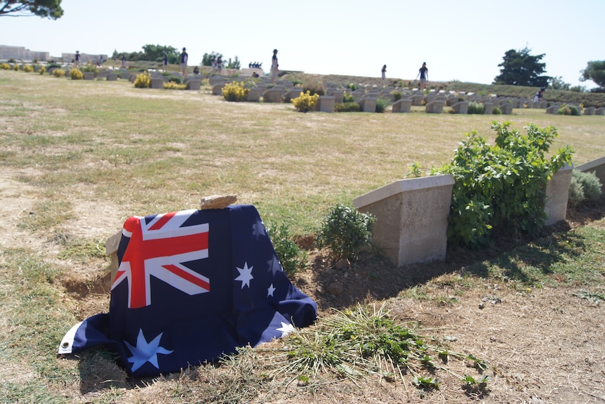 An Australian flag is draped over a short headstone in Lone Pine Cemetery in Gallipoli. The grass around it is dry