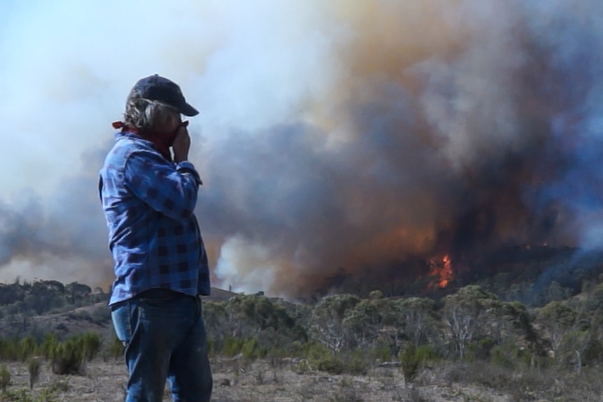 A man covers his mouth to reduce smoke inhalation as he stands in front of a bush fire