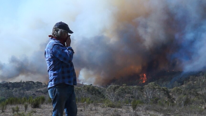 A man covers his mouth to reduce smoke inhalation as he stands in front of a bush fire