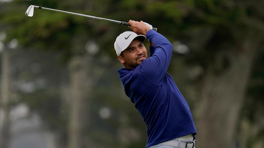 Jason Day swings his golf club wearing a blue jumper with a smile on his face