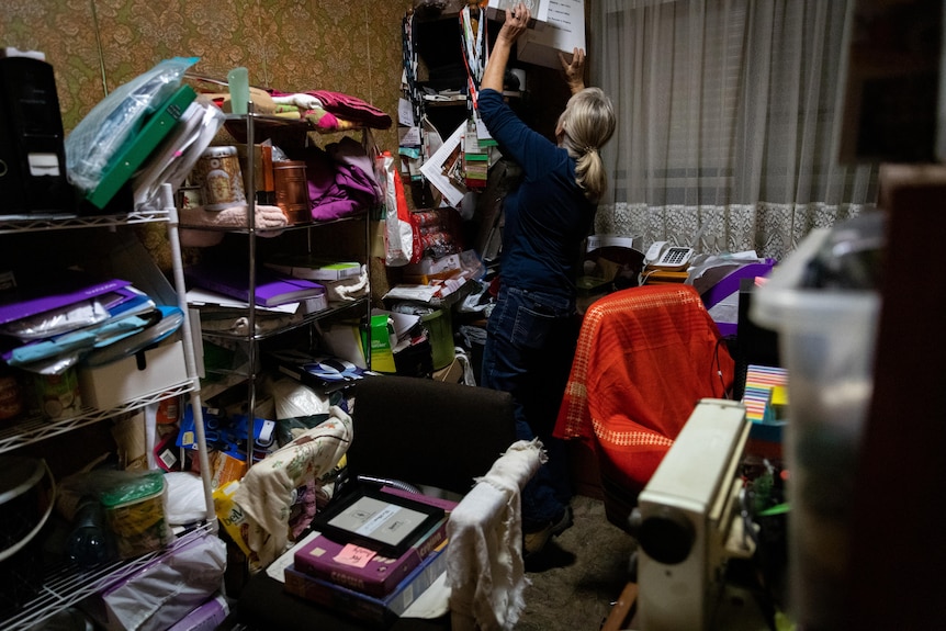 A woman sifts through boxes of belongings in a small cluttered room.