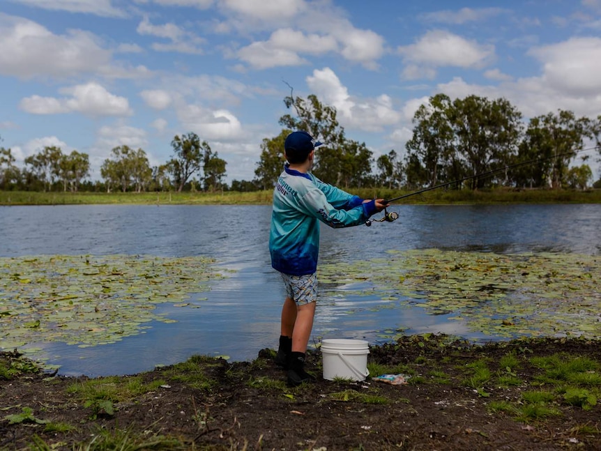 A young boy with a fishing rod, stands with back to camera, casts a line into a lake with lilies. Blue sky, white clouds.