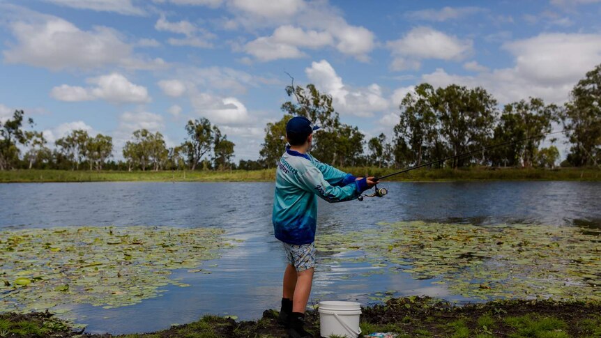 A young boy with a fishing rod, stands with back to camera, casts a line into a lake with lilies. Blue sky, white clouds.