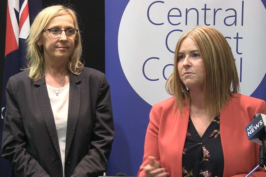 Two women with blonde hair stand on a stage, giving an address.