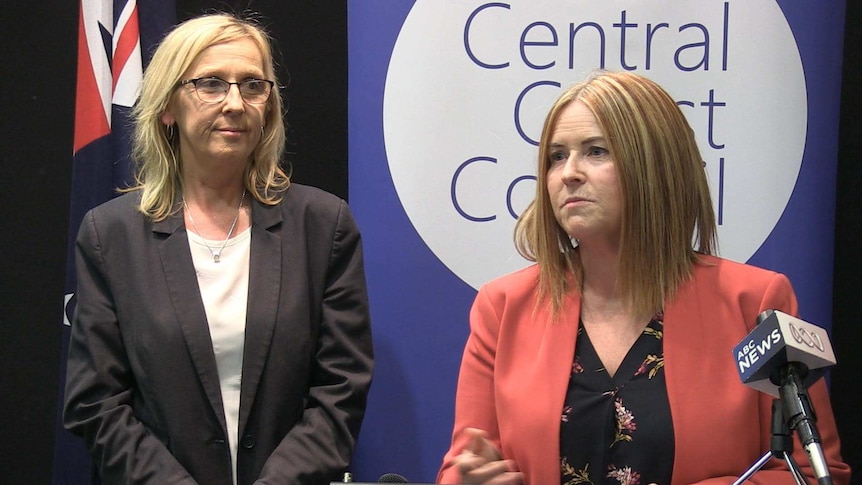 Two women with blonde hair stand on a stage, giving an address.