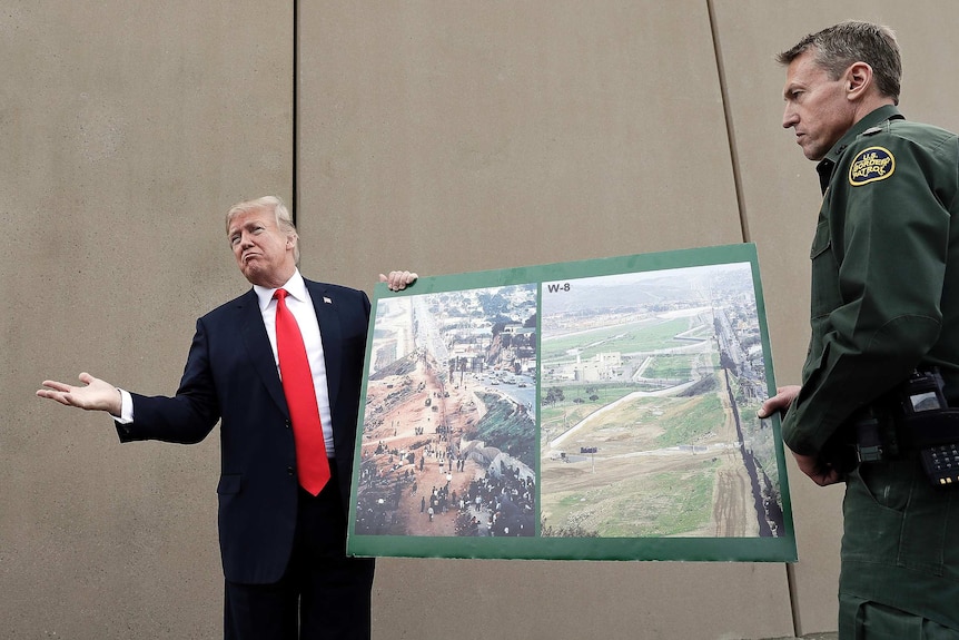 Trump stands in a suit wearing red tie and gestures while holding a large aerial map of wall with a border patrol official.