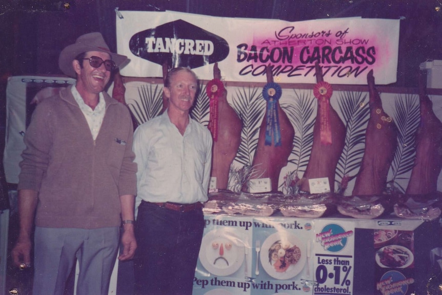 Two men standing in front of a row of bacon carcases with ribbons indicating the places earned in a show competition