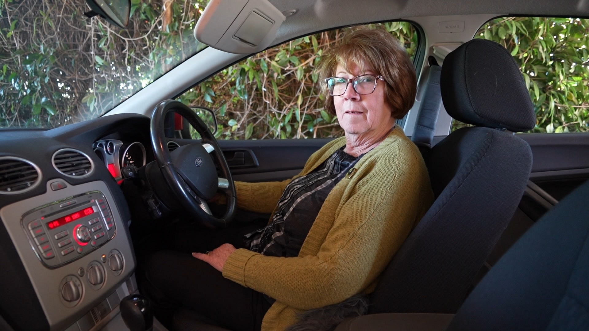 Elderly woman sitting in driver's seat of her small car, green bushes outside her window