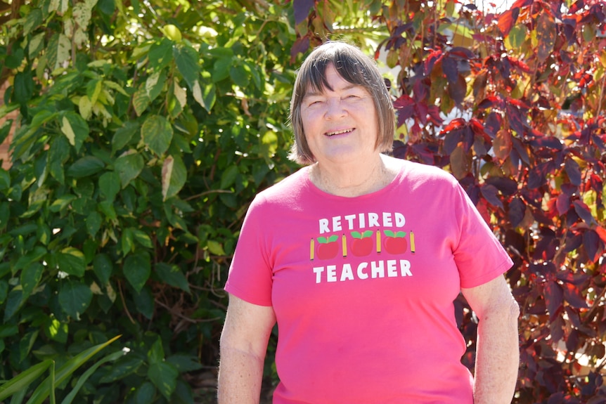 Older woman wearing a pink shirt that says 'Retired Teacher' standing in front of trees.