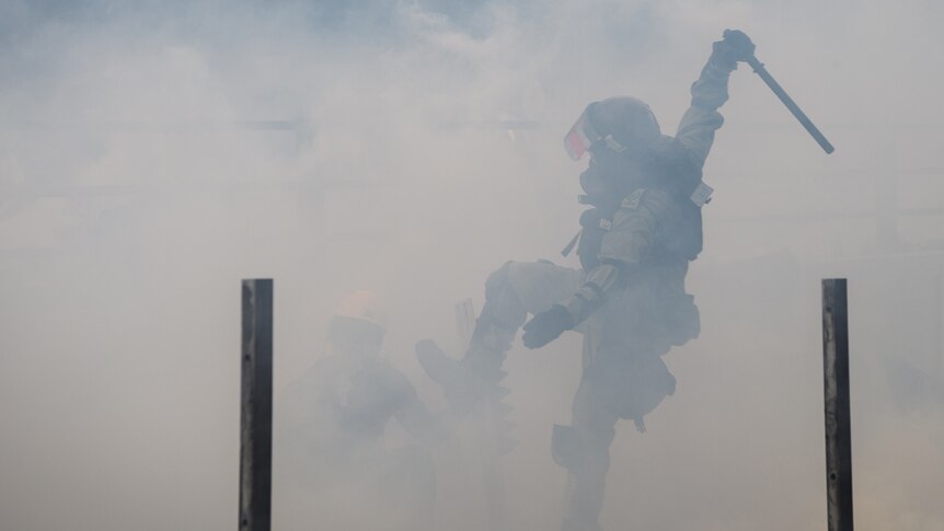 A Hong Kong police officer is viewed through a cloud of tear gas as they raise their baton and leg with a protester crouching.