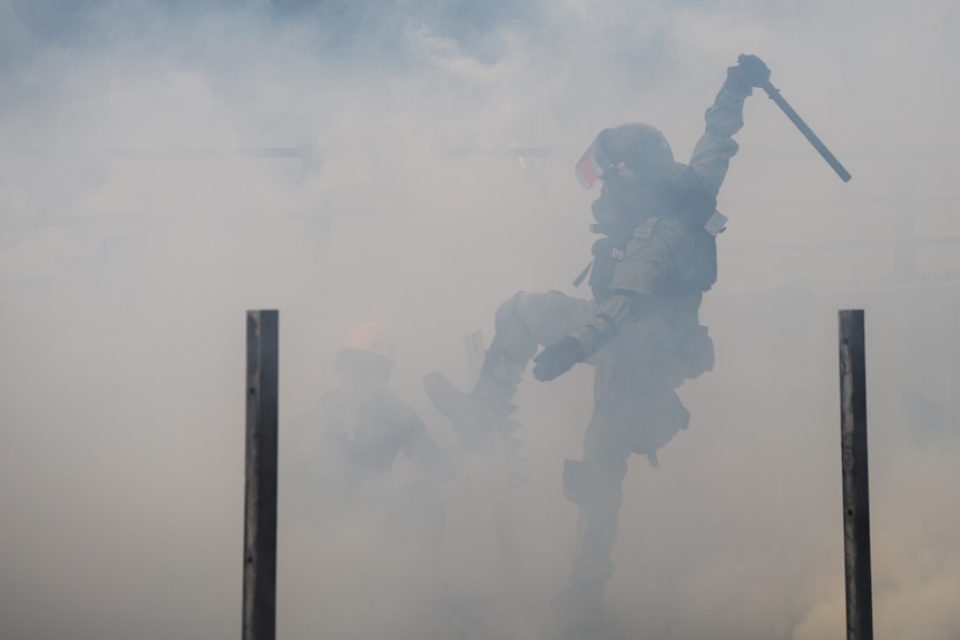 A Hong Kong police officer is viewed through a cloud of tear gas as they raise their baton and leg with a protester crouching.