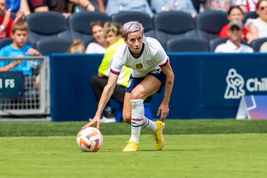 Megan Rapinoe kneeling down to reset a ball on a soccer pitch at a game