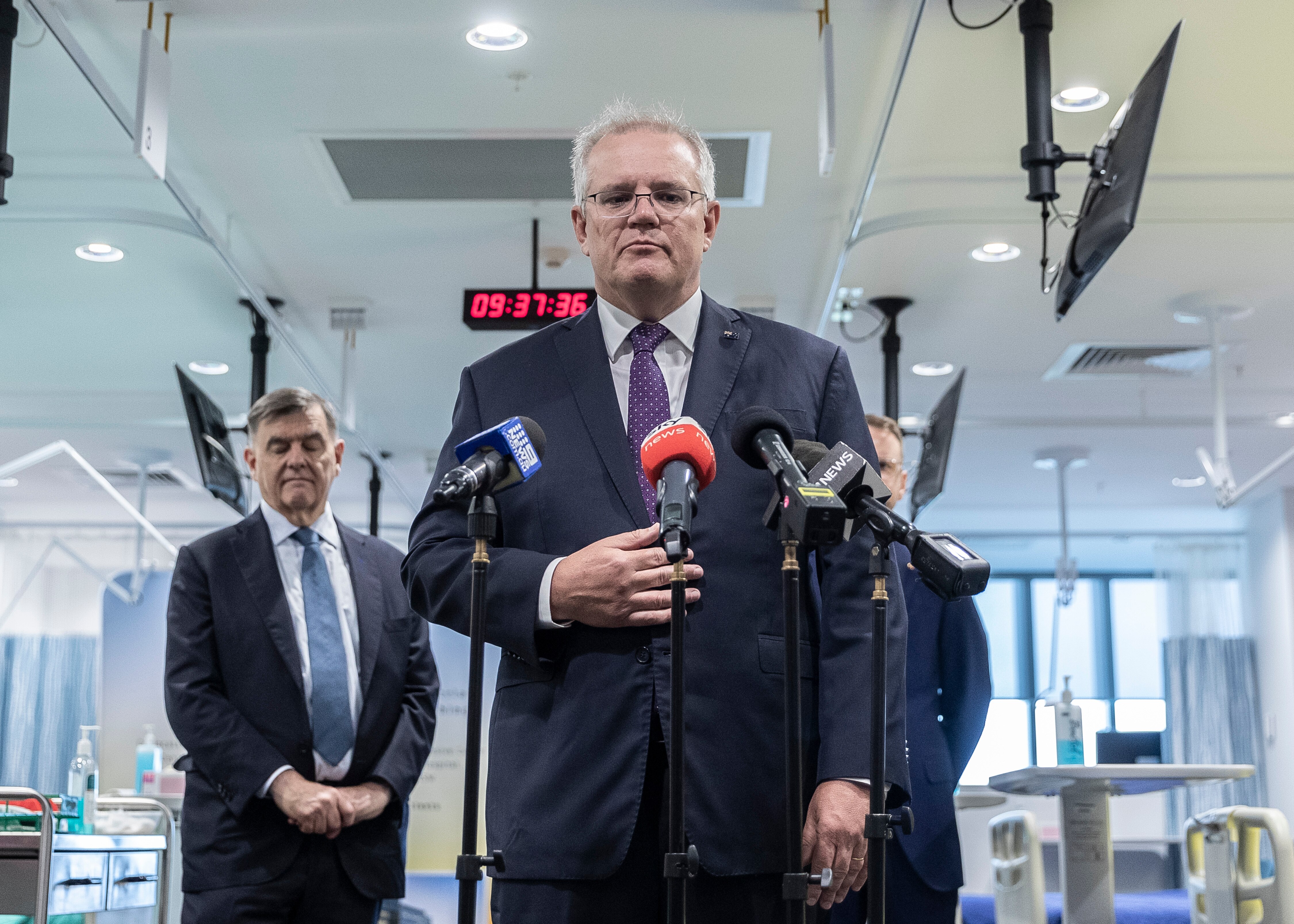 A frowning Scott Morrison stands in front of microphones in a hospital room. Next to him is Brendan Murphy.