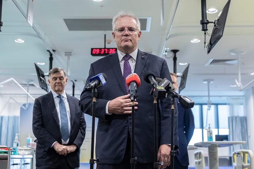 A frowning Scott Morrison stands in front of microphones in a hospital room. Next to him is Brendan Murphy.