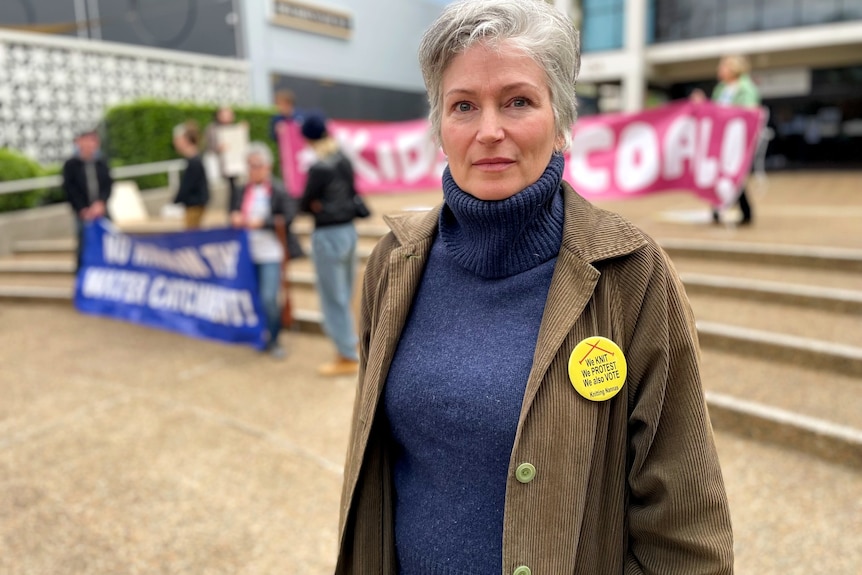 Woman with short grey hair a coat and badge looks seriously at the camera, people hold banners in the background.