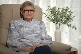 A woman with glasses sitting in an armchair next to a plant