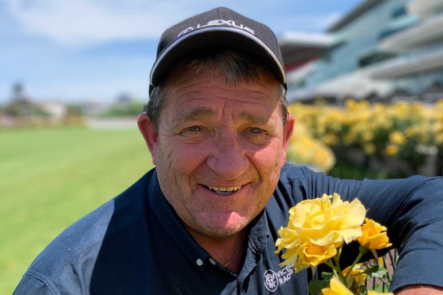 A smiling man in a baseball cap on a racecourse posing with yellow roses.