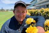 A smiling man in a baseball cap on a racecourse posing with yellow roses.