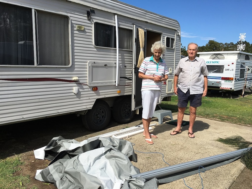 Elderly man and woman stand in front of caravan with annexe strewn on the ground in front of them.
