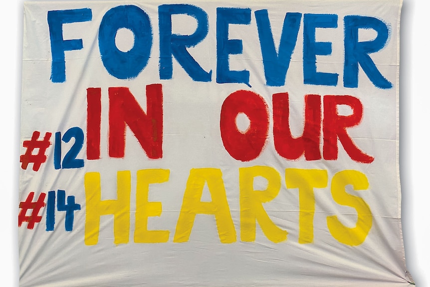 A hand painted flag in Crows team colours , blue, red, gold, reads "Forever in our hearts", player number 12 and number 14