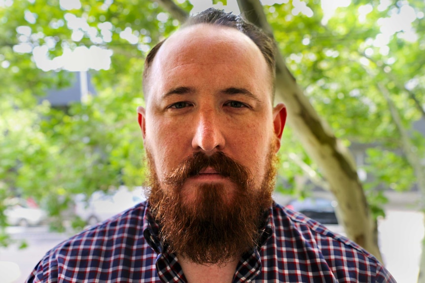 Man with think brown beard stares into camera with tree lined street behind.