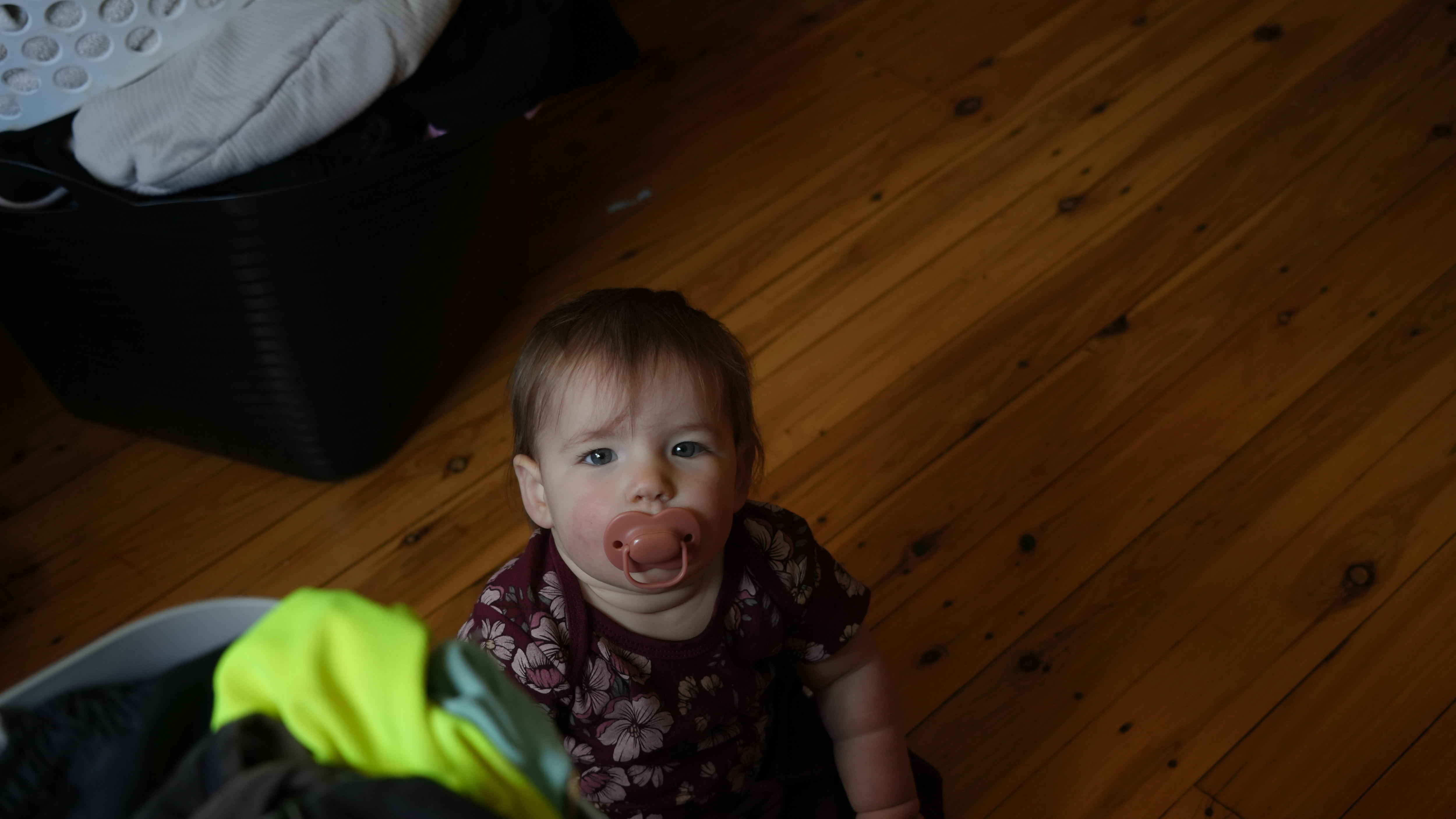 A toddler sucks a dummy while sitting on the floor looking up at the camera.