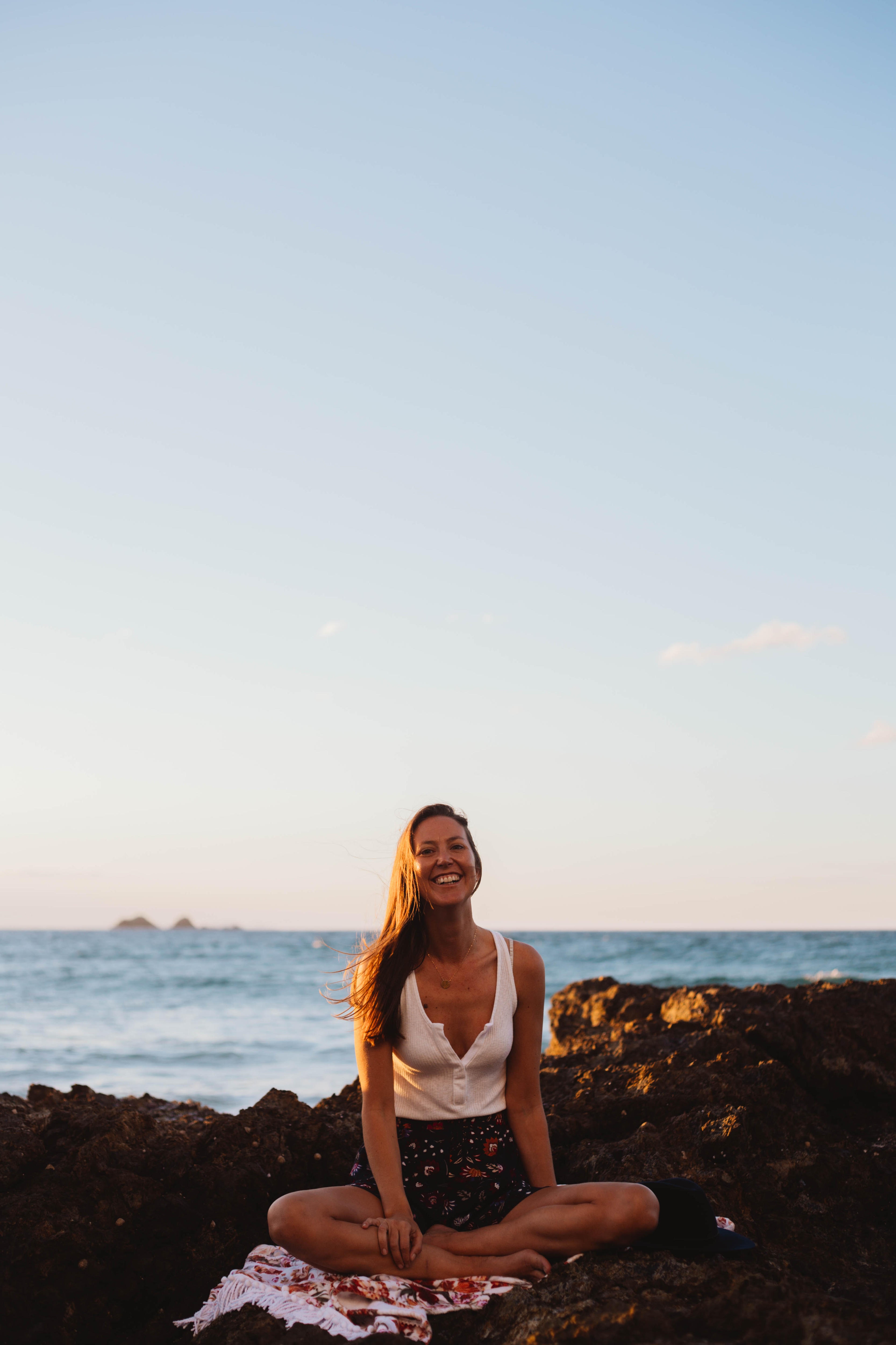 A woman sits cross legged on the beach with a big smile. 