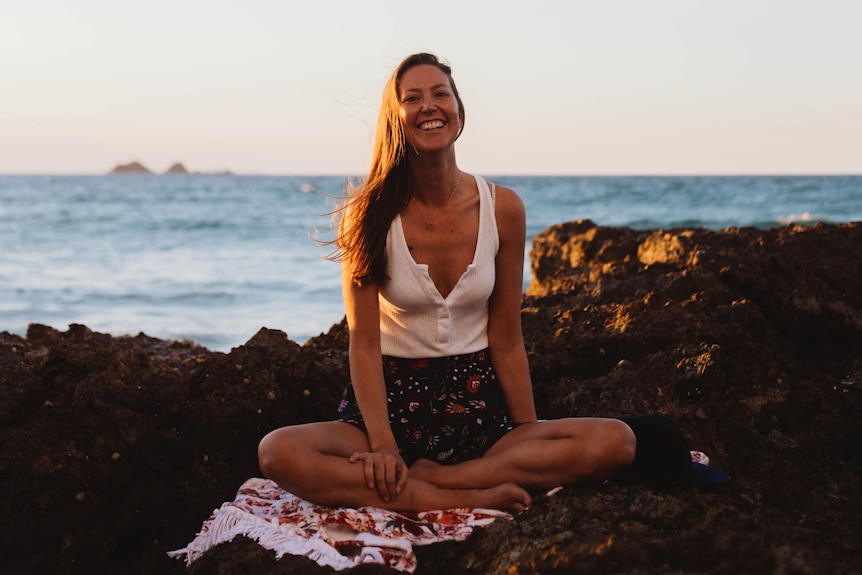 A woman sits cross legged on the beach with a big smile. 