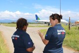 Two women stand in AUSAID polo shirts in front of a plane.