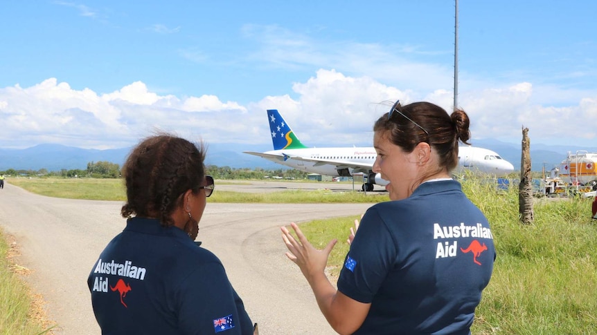Two women stand in AUSAID polo shirts in front of a plane.