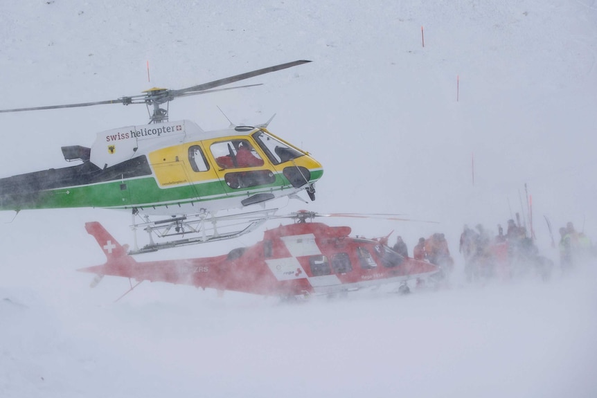 A yellow and green helicopter tried to land in heavy snow. A red one is already on the ground with rescue workers around it.