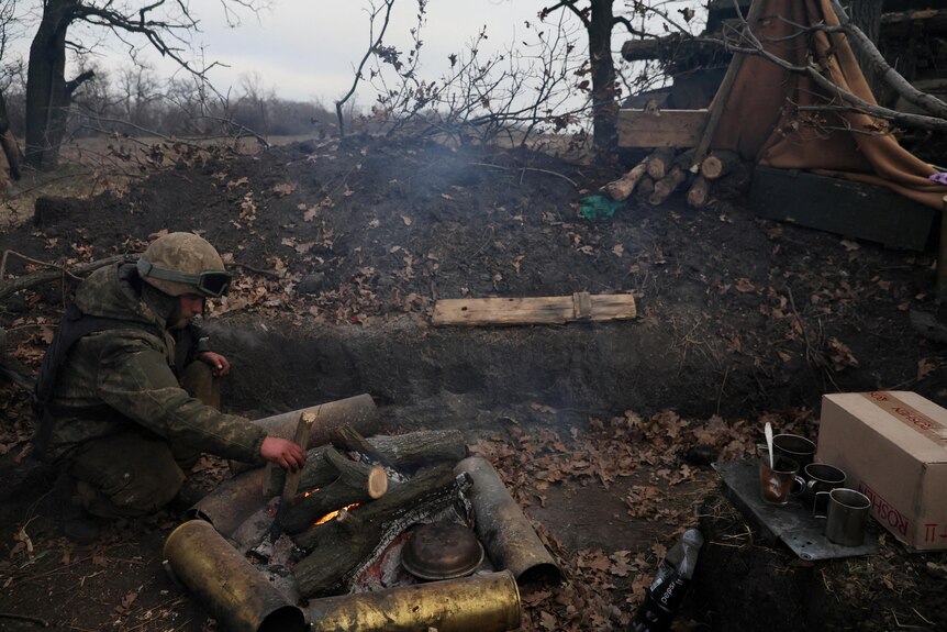 A soldier adds wood to a campfire while sitting in a trench