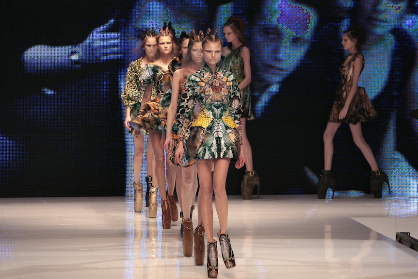 Runway shot showing line of models in structured couture garments in reptile patterns. 