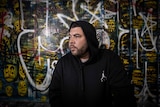 A man in a black hoodie looks to the left as he poses in front of a graffiti wall
