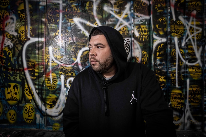 A man in a black hoodie looks to the left as he poses in front of a graffiti wall