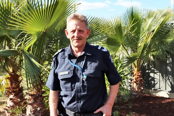 A middle aged man with a moustache and a police shirt on stands smiling in front of trees.