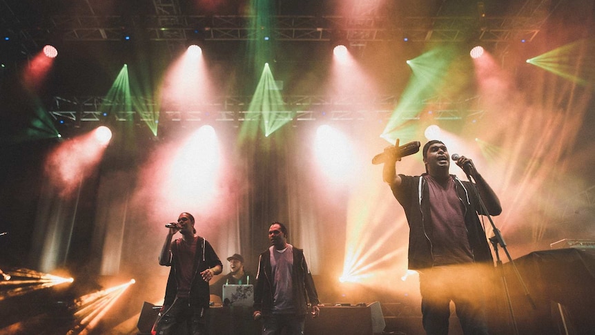 Indigenous performers on music festival stage with bright lights.