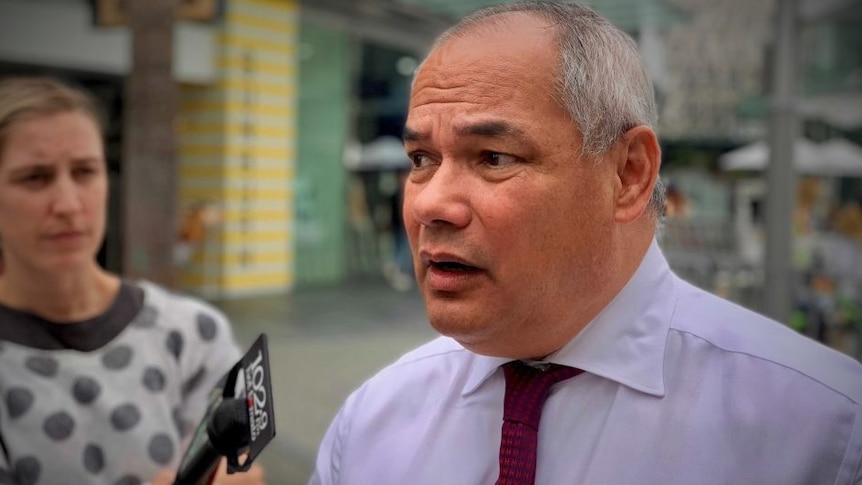 A man in a blue shirt and tie talks into a microphone at an outdoor press conference.