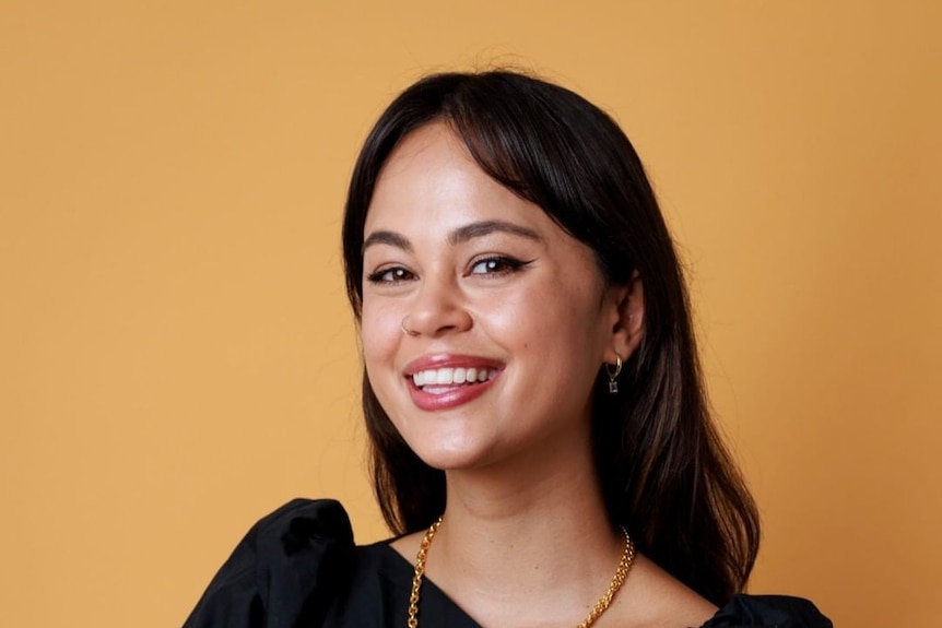 A woman in her 20s smiles with an orange backdrop, wearing a black dress 