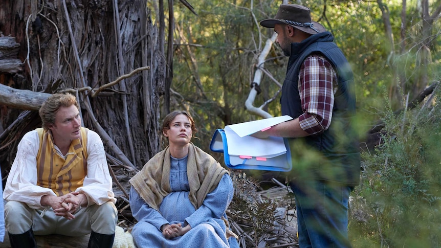 A male and female actor in clothing from the colonial era look up at the script writer during an Australian bush shoot.