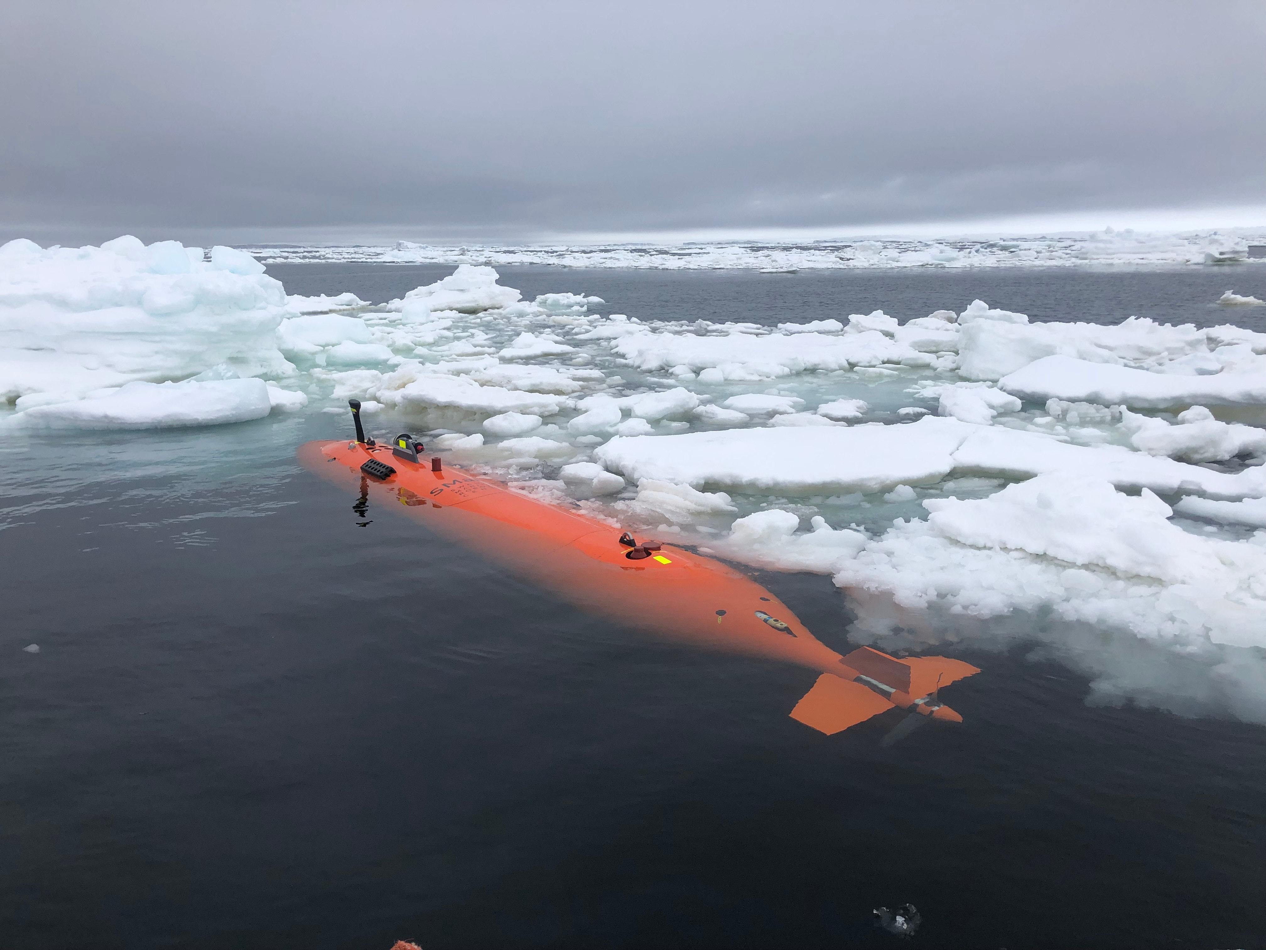 A bright orange cylindrical unmanned submarine floats amongst sea ice on the surface of dark, deep water.