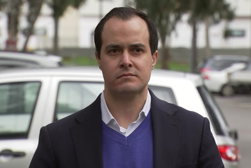 A man wearing a black suit jacket, blue jumper and white shirt stands in a car park with a serious expression