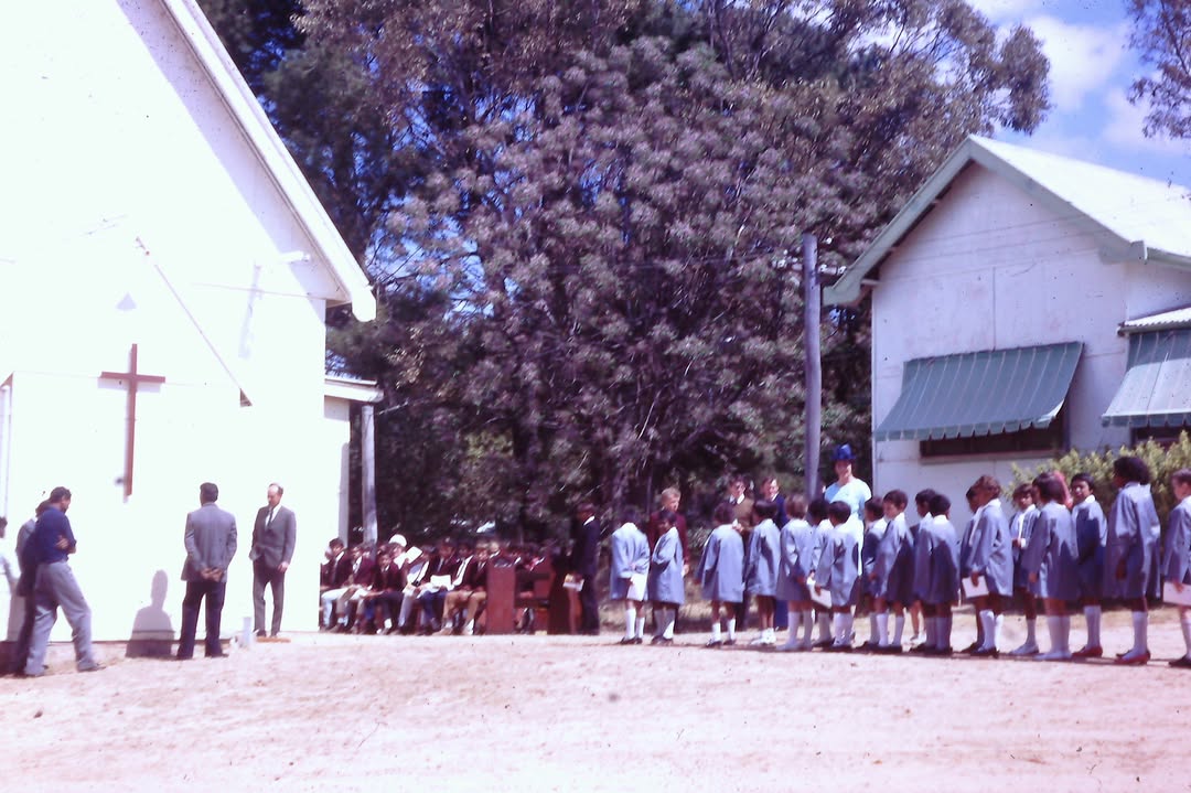 Una iglesia blanca con una cruz, niños alineados con batas y otros sentados en bancos afuera.