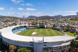 A shot looking over the stands and into a cricket ground, with four lighttowers and the city of Hobart in the backgro