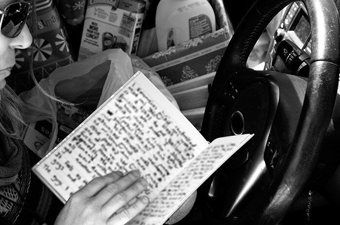 A black and white photo of a woman, with only the edge of her face shown, holding a notepad while sitting in a car.