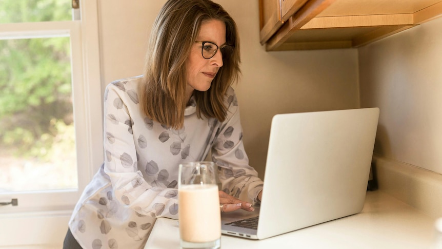 Woman with long hair and glasses leaning against kitchen bench looking at her laptop with a glass of milk next to it